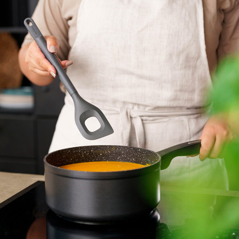 The Zyliss Angled Mixing Spoon is a gray silicone utensil with an ergonomic handle and a rectangular hole in the head, designed for non-stick cookware. Shown on a plain white background. 