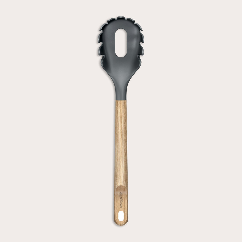 The Zyliss Oak Wood Spaghetti Server is a black utensil with an oak handle, pronged edges, and a large oval center hole, shown on a plain light background.