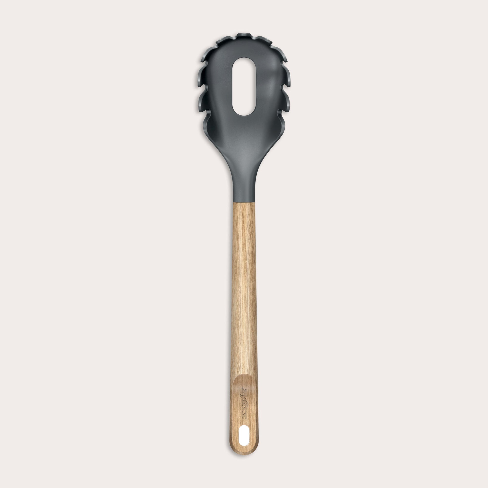 The Zyliss Oak Wood Spaghetti Server is a black utensil with an oak handle, pronged edges, and a large oval center hole, shown on a plain light background.