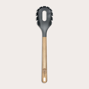 The Zyliss Oak Wood Spaghetti Server is a black utensil with an oak handle, pronged edges, and a large oval center hole, shown on a plain light background.
