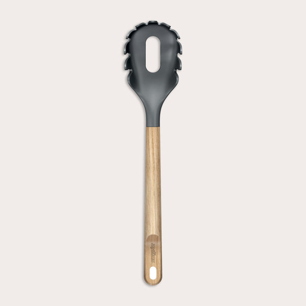 The Zyliss Oak Wood Spaghetti Server is a black utensil with an oak handle, pronged edges, and a large oval center hole, shown on a plain light background.