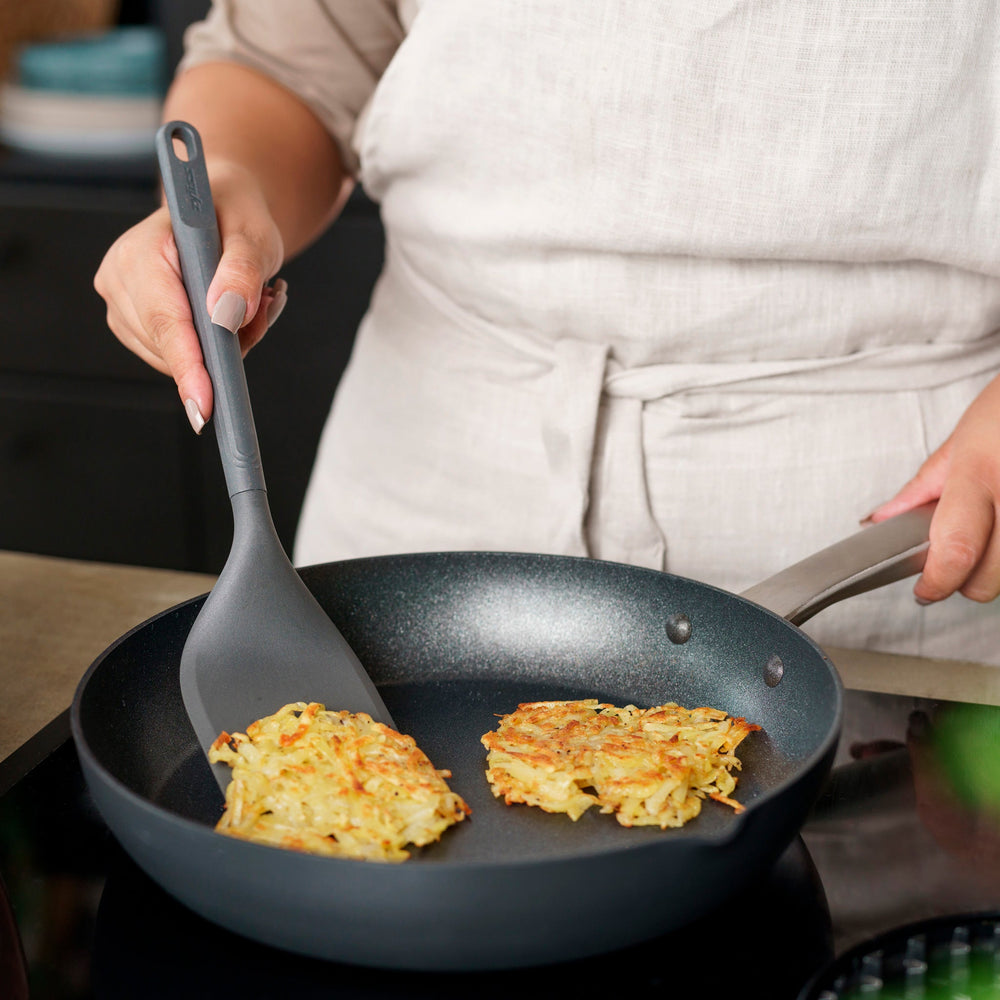 The Zyliss Angled Turner is a dark gray, heat-resistant silicone utensil with a slightly angled rectangular head and long handle, shown on a plain light background. Safe for use on non-stick surfaces. 