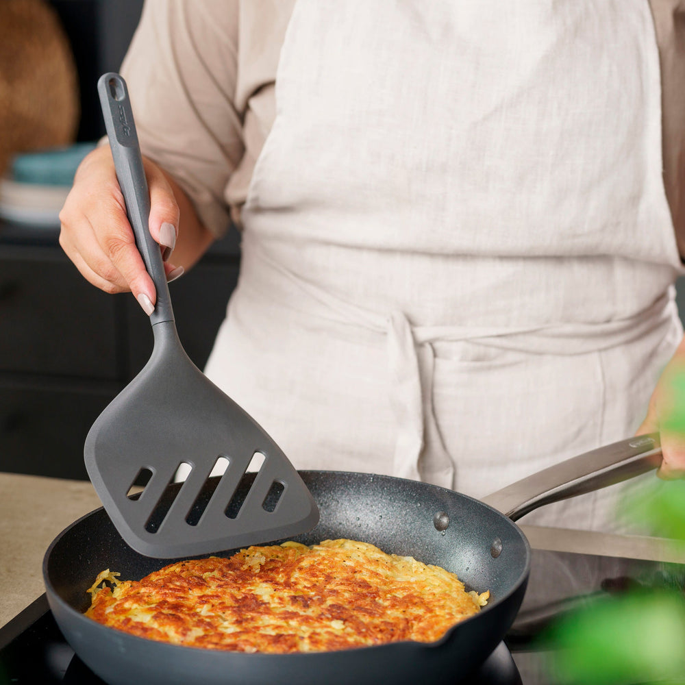 A Zyliss Turner XL slotted spatula in black, featuring a wide square head with three diagonal slots, ergonomic handle, and heat-resistant design is displayed against a plain light background. 