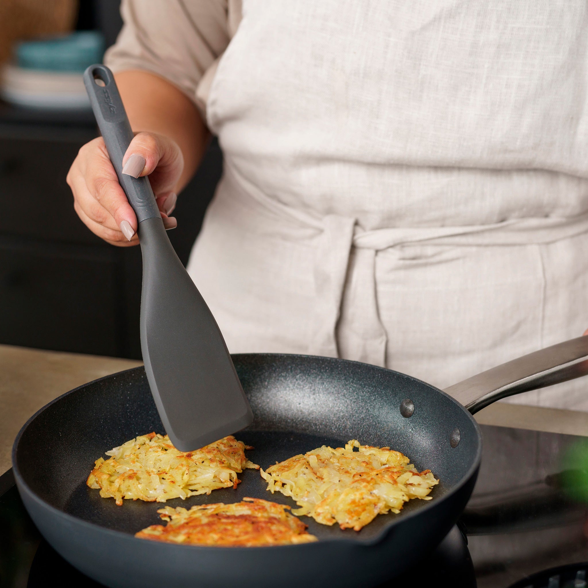 Wearing a light apron, someone uses the Zyliss Easy Lift Turner to cook three potato pancakes in a nonstick frying pan on the stove. 