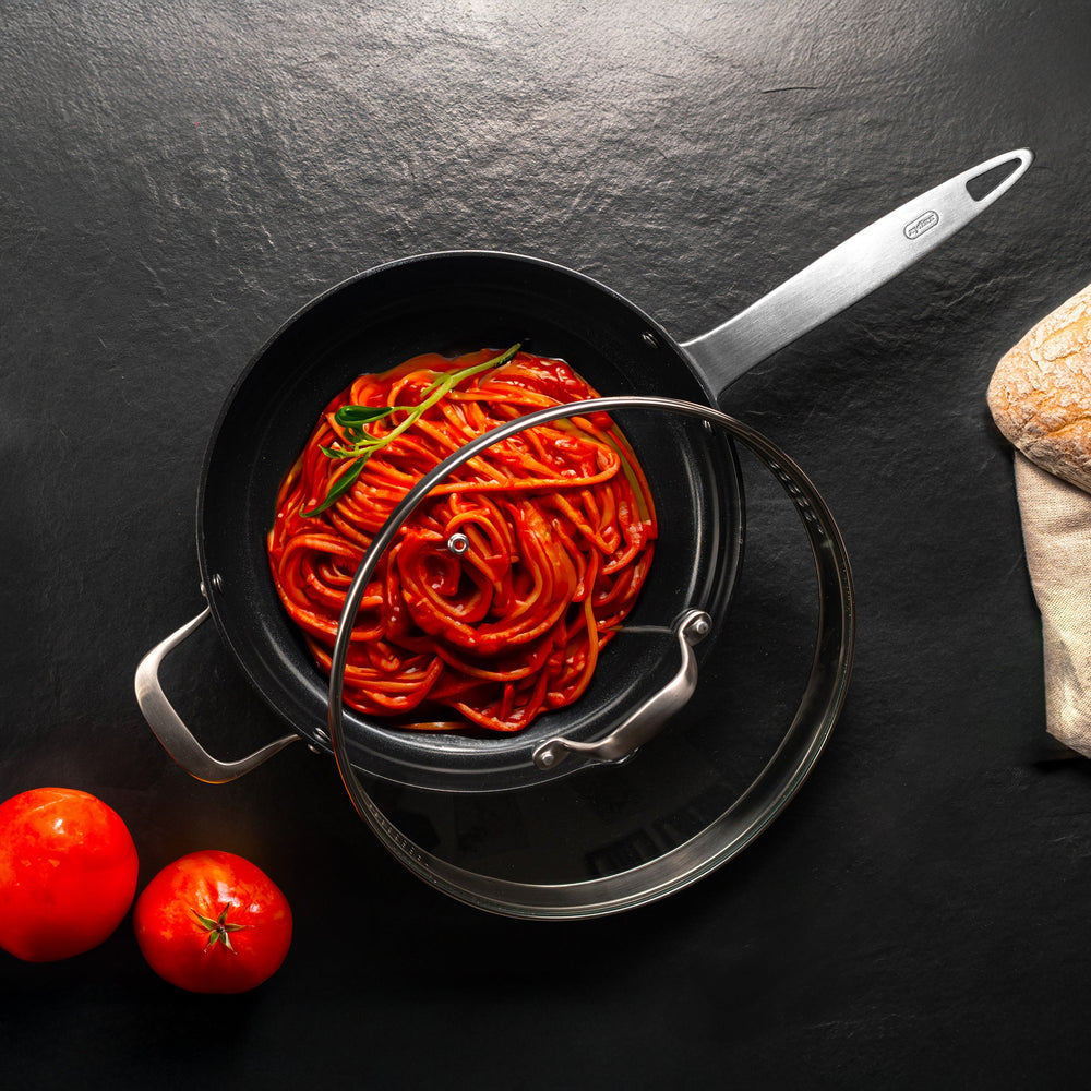 A Zyliss Ultimate Pro Ceramic Saute Pan With Lid, featuring a black nonstick surface, glass lid, long metal handle, and short helper handle, is displayed against a plain light background. 