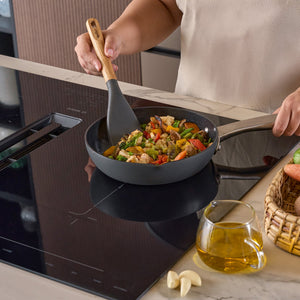 A person cooks a colorful vegetable stir-fry in a black pan on an induction cooktop, using the Zyliss Oak Wood Easy Lift Turner. Nearby on the counter are a small pitcher of oil, garlic cloves, and a basket of vegetables.
