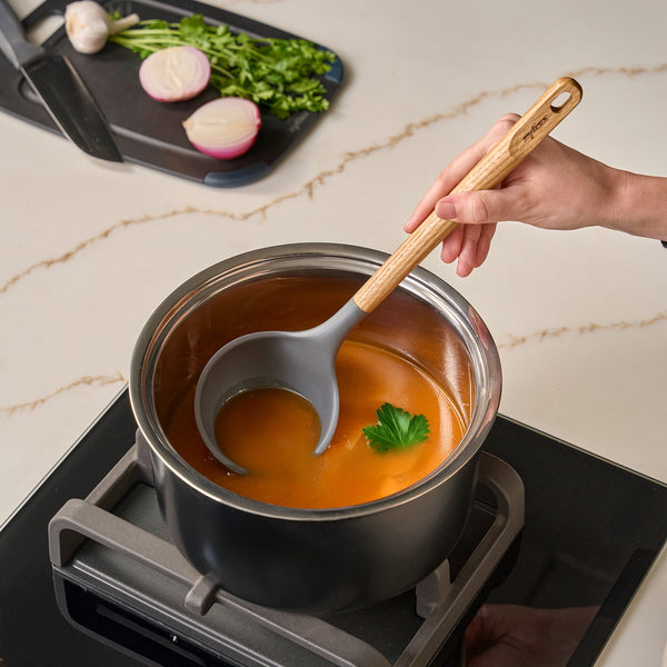 A hand holds the Zyliss Oak Wood Ladle Large above a pot of orange soup with parsley, on a stovetop. In the background, a cutting board displays onions, garlic, and fresh herbs.