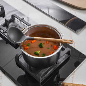 A pot of soup with carrots and broccoli simmers on the stove, while a Zyliss Oak Wood Ladle Large rests across the top. A slice of bread is partially visible in the foreground.
