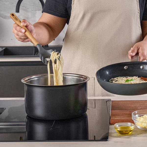 Wearing a beige apron, a person uses the Zyliss Oak Wood Spaghetti Server to lift cooked spaghetti from a pot on the stove, with a nearby pan and small ingredient bowls.