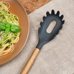The Zyliss Oak Wood Spaghetti Server with a black finish rests on a beige cloth next to a plate of spaghetti garnished with green pesto and fresh basil.