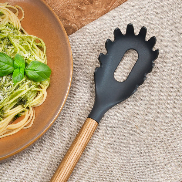 The Zyliss Oak Wood Spaghetti Server with a black finish rests on a beige cloth next to a plate of spaghetti garnished with green pesto and fresh basil.