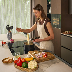 In a modern kitchen with oak cabinets, a woman in an apron cooks pasta while using the Zyliss Oak Wood Spaghetti Server. Fresh vegetables, bread, and white wine are arranged on the counter in front of her.