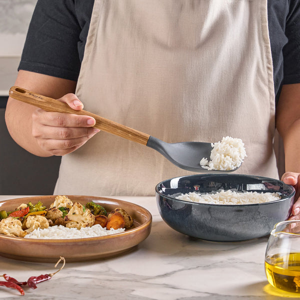 Wearing a beige apron, someone serves white rice into a dark bowl with the Zyliss Oak Wood Spoon Large. Nearby, there’s a plate of rice and vegetables and a small glass oil container on the counter.