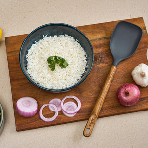 A bowl of cooked white rice with cilantro rests on a wooden board beside a Zyliss Oak Wood Spoon Large, sliced red onion, a whole onion, and a garlic bulb.