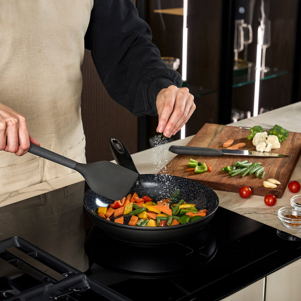 #24cm/9½"
Wearing an apron, a person sprinkles salt over vegetables cooking in a Zyliss Ultimate Ceramic Fry Pan on the stove, holding a spatula, with extra veggies on a cutting board in the background.