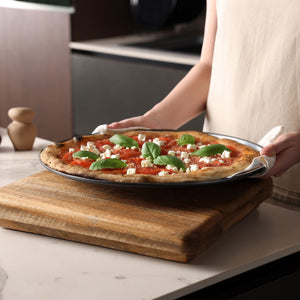 A person holds a Zyliss Non-Stick Pizza Tray with a freshly baked pizza topped with tomato sauce, white cheese, and basil over a wooden board—ideal for enjoying crispy pizza bases in the kitchen.
