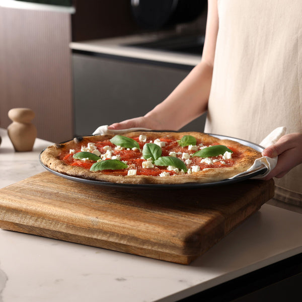 A person holds a Zyliss Non-Stick Pizza Tray with a freshly baked pizza topped with tomato sauce, white cheese, and basil over a wooden board—ideal for enjoying crispy pizza bases in the kitchen.