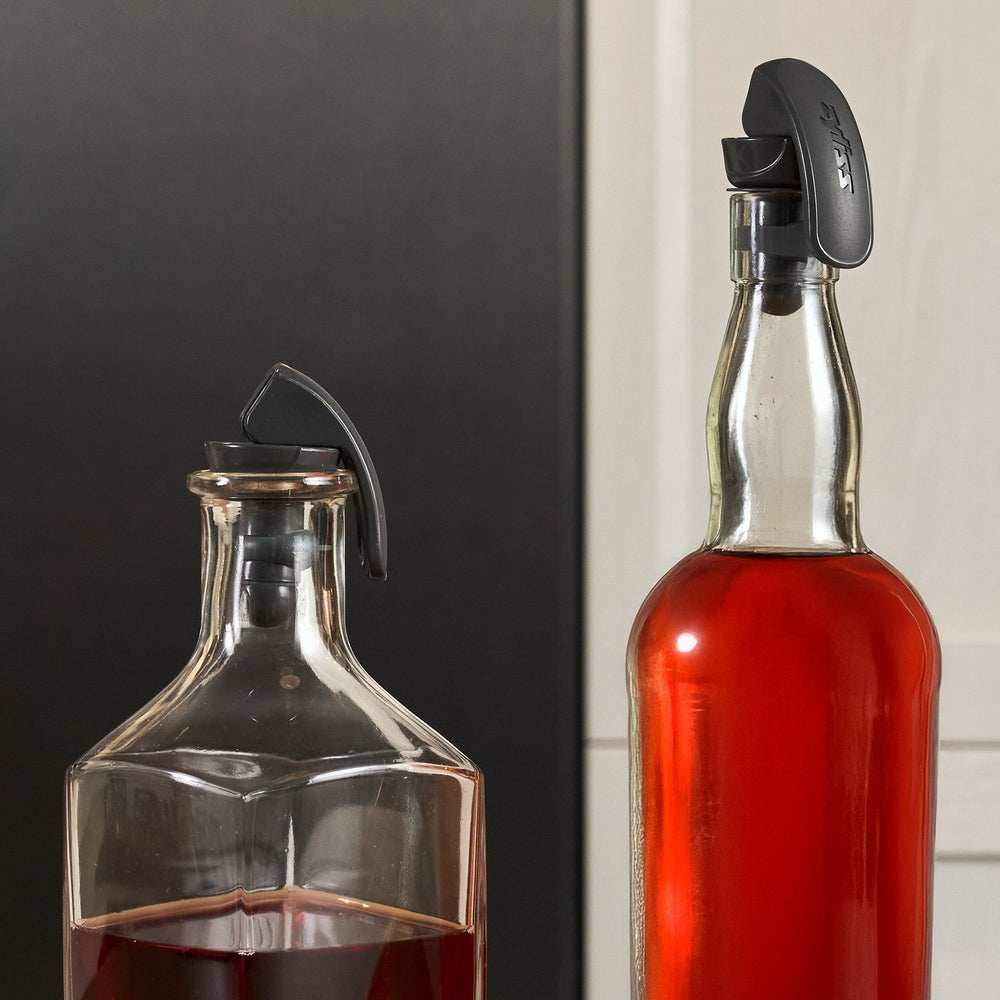 Two black, curved Zyliss Bottle Stopper 2 Piece Set items with a matte finish are displayed upright on a light beige background, featuring sleek designs that resemble wireless earbuds and small brand markings on their surfaces.