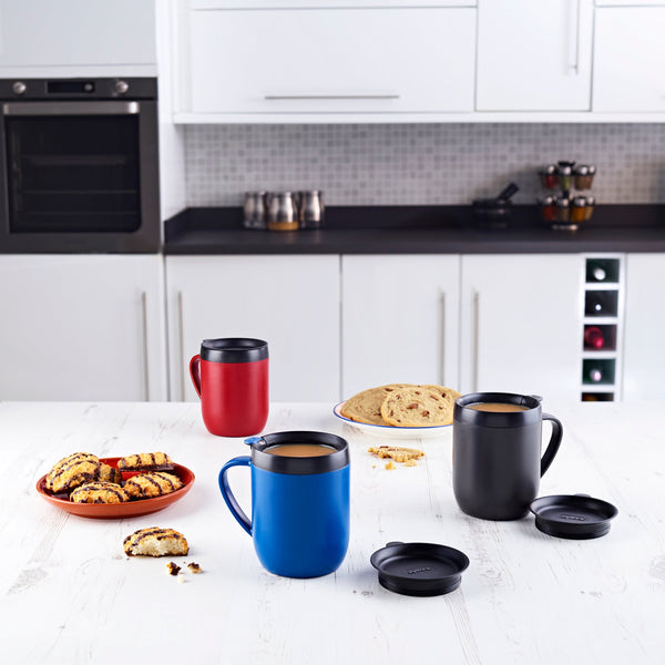 A kitchen scene with a white countertop displaying three travel mugs (red, blue, and black), plates of assorted cookies, and removable mug lids, set against white cabinets and built-in ovens.