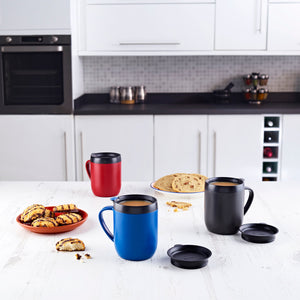Three Zyliss Hot Mug Cafetière Mugs in blue, red, and black with double-wall insulation sit on a white kitchen table with cookies and bread. Two black lids are nearby. Modern white cabinets and appliances complete this coffee lovers scene. #grey