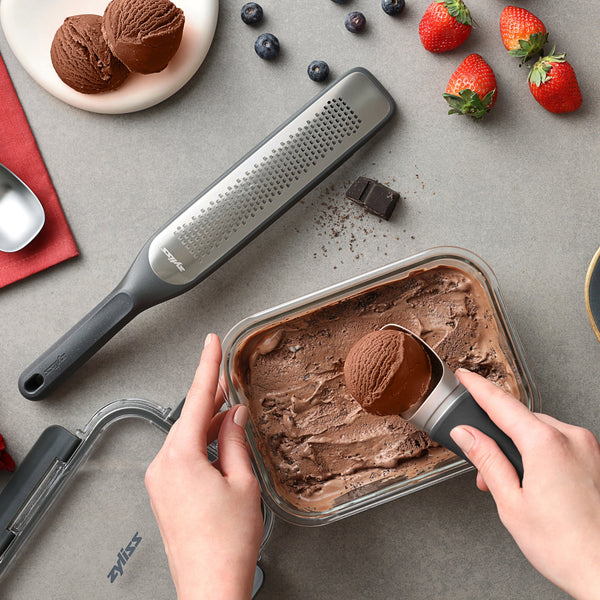 #0.64L/0.67qt A person scoops chocolate ice cream from a rectangular Zyliss Glass Container. Nearby are a grater, chocolate pieces, blueberries, strawberries, an ice cream scoop, and a red napkin on a gray surface—ideal for easy meal prep or dessert storage!.