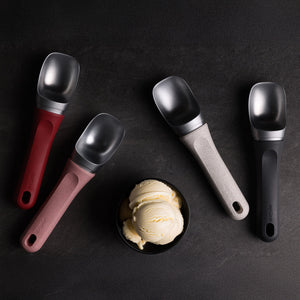 A bowl of vanilla ice cream rests on a dark surface, encircled by four Zyliss Ice Cream Scoops with eco-friendly red, pink, gray, and black handles—each neatly arranged and dishwasher-safe for easy cleaning.