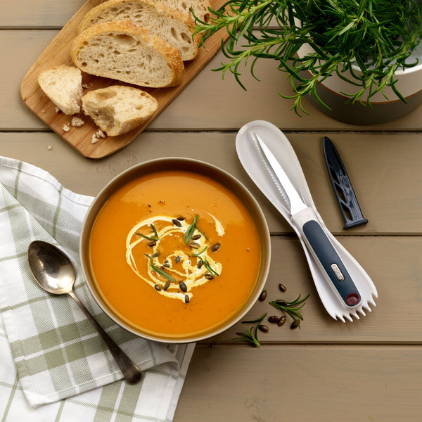 A bowl of creamy orange soup sits on a wooden table, garnished with cream, seeds, and herbs. Its made using the Zyliss 3 In 1 Pumpkin & Squash Scoop & Knife. Nearby are bread slices, a serrated knife, cutlery, napkin, and potted rosemary. 