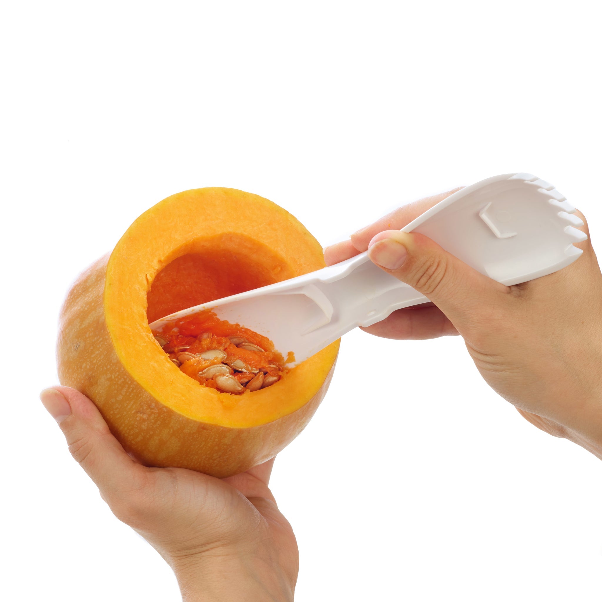 A person uses a white plastic tool to scoop seeds and pulp from the inside of a hollowed-out pumpkin against a white background.