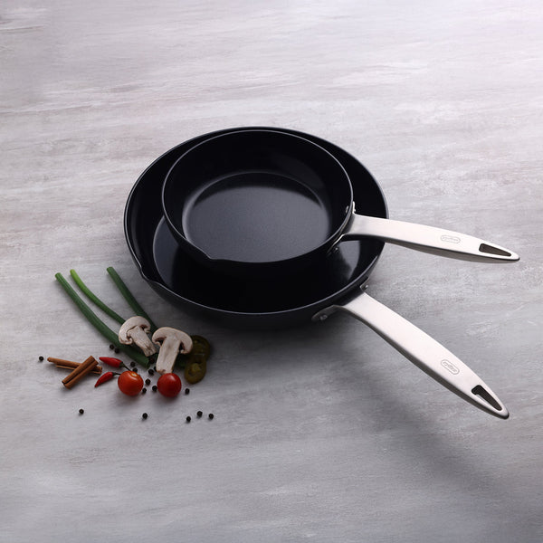Two Zyliss Ultimate Pro Ceramic Fry Pans, black with silver handles and made from recycled aluminum with a non-stick coating, are stacked on a light gray surface alongside green onions, mushrooms, cherry tomatoes, chili, and spices. #20cm/8"
