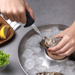 Using the Zyliss Oyster Knife Shucker Paddy, someone expertly shucks oysters over a bowl of ice. A lemon wedge sits on a wooden tray with lettuce nearby on the table.