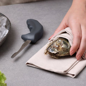 A person holds an oyster in a cloth, ready to open it with the Zyliss Oyster Knife Shucker Paddy on a gray surface; a bowl of ice sits nearby for fresh oyster shucking.