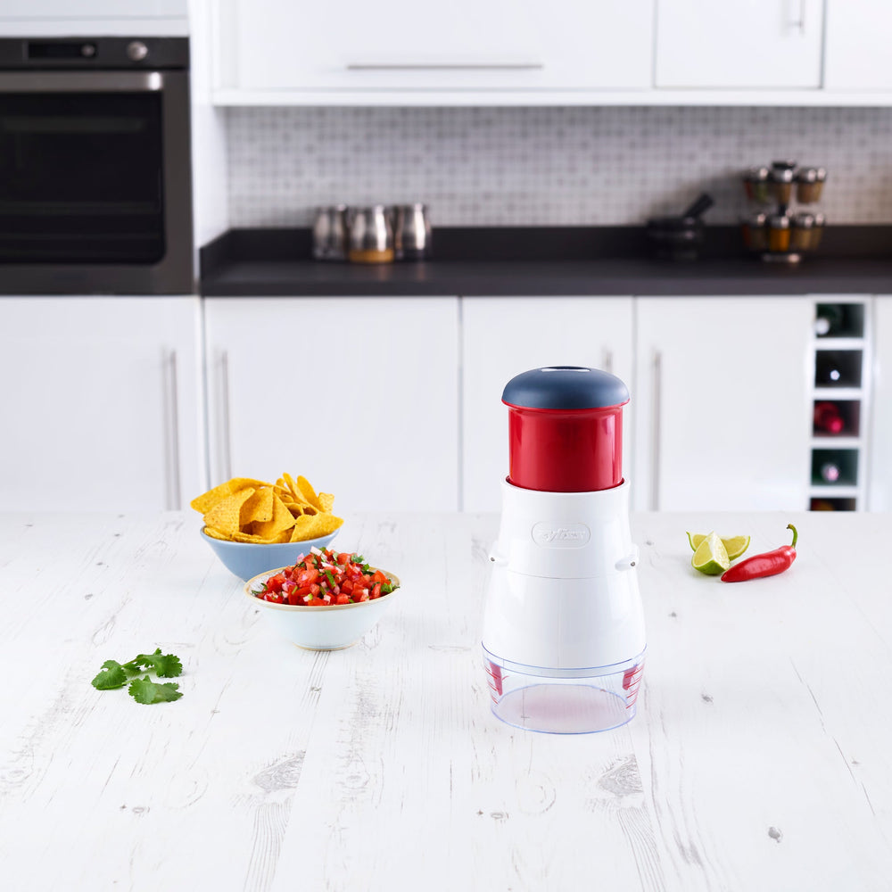 The Zyliss Zick-Zick 2 Food Chopper stands upright on a clear and black base, featuring a black top, red middle section, and white body, resembling a storage container against a plain light background. 