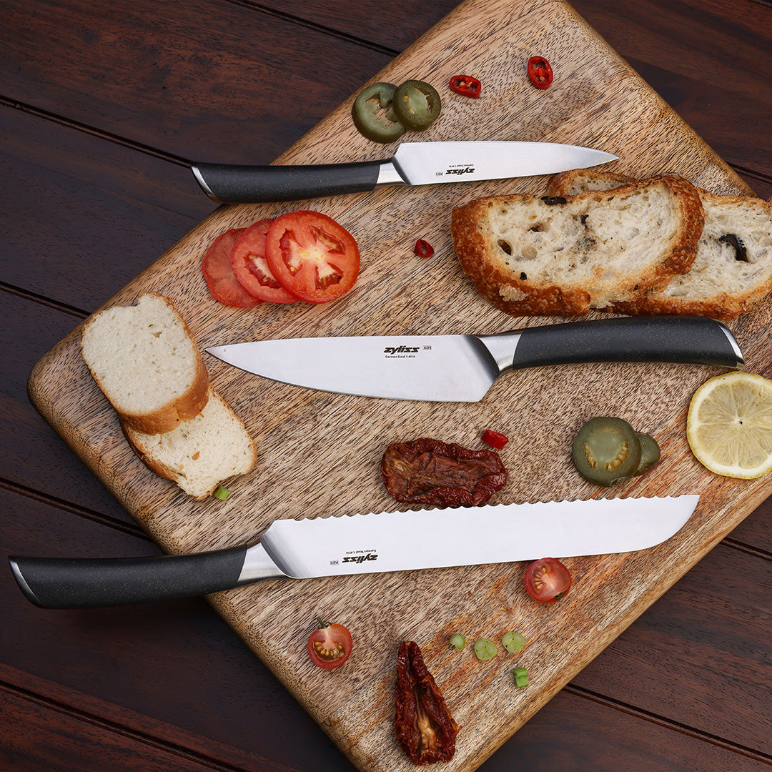 The Zyliss Comfort Pro Chef + Santoku Knife Set, featuring three kitchen knives with black handles and stainless steel blades, rests on a wooden cutting board surrounded by sliced bread, tomato, lemon, pickles, chili, and sun-dried tomatoes. The board is placed on a dark wooden surface.