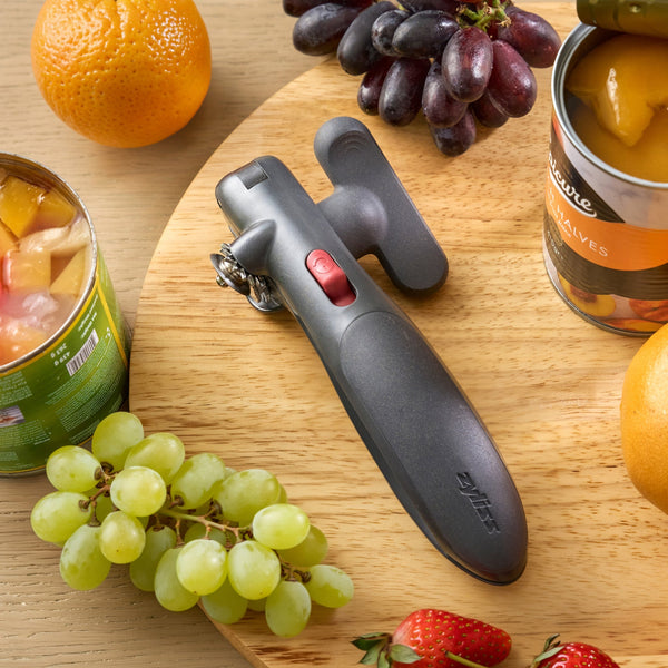 A black manual can opener with a red switch, displayed on a wooden board surrounded by fruit including grapes, strawberries, oranges, and canned fruit.