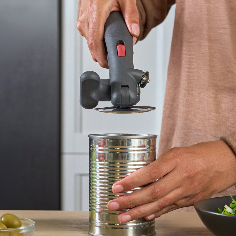 A person using a manual can opener to open a metal can on a kitchen counter, with canned olives, salad, and other kitchen items nearby.
