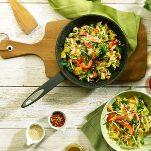 The Zyliss Ultimate Non-Stick Frying Pan, filled with vibrant stir-fried veggies, rests on a wooden board beside a matching bowl. Nearby, small bowls of sesame seeds and chili flakes sit on a rustic white table. #20cm/8"&28cm/11"