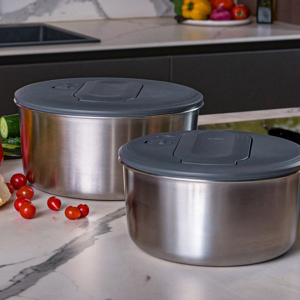 The Zyliss Swift Dry Salad Spinner Stainless Steel, with dark gray lids, sits on a kitchen countertop surrounded by fresh cherry tomatoes and cucumbers—ideal for prepping crisp salads.