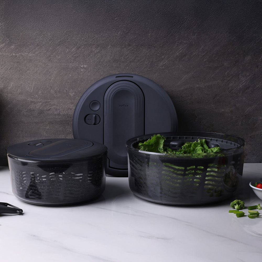 The Zyliss Swift Dry Salad Spinner features a transparent, round bowl with a dark gray lid and one-handed pump handle. Its slitted inner basket is visible through the clear outer bowl, shown on a light background.