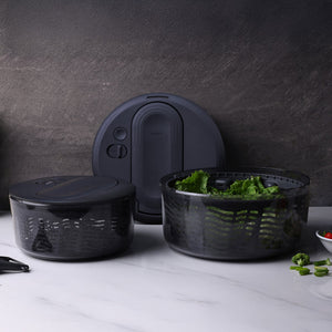 The Zyliss Swift Dry Salad Spinner, featuring a black lidded bowl and strainer basket, rests on a white countertop filled with greens. Its one-handed pump design stands out against a textured dark gray background.