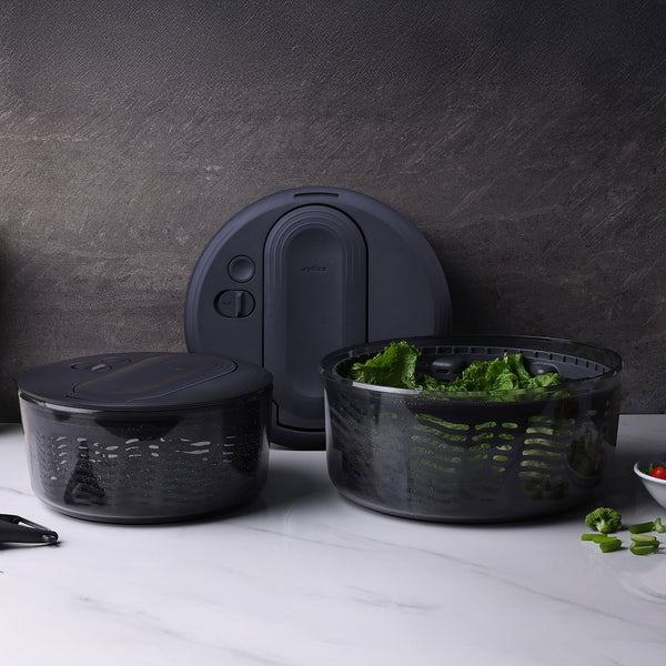 The Zyliss Swift Dry Salad Spinner, featuring a black lidded bowl and strainer basket, rests on a white countertop filled with greens. Its one-handed pump design stands out against a textured dark gray background.