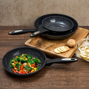 Three Zyliss Ultimate Ceramic Fry Pans, one filled with sautéed vegetables, are set on a wooden table with a cutting board, sliced potatoes, whole potatoes, and a bowl of potato slices—showcasing stylish PFAS-free cookware.