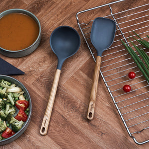 A Zyliss Oak Wood Spoon Large rests on a wooden surface beside a bowl of soup, a salad, and a cooling rack topped with green onions and cherry tomatoes.