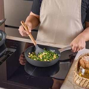 A person in a beige apron cooks corn and peas in a frying pan on an induction stove, stirring with a Zyliss Oak Wood Spoon Large. A basket of bread and a glass of oil sit on the counter nearby.