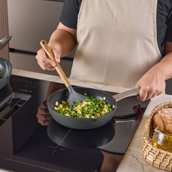 A person in a beige apron cooks corn and peas in a frying pan on an induction stove, stirring with a Zyliss Oak Wood Spoon Large. A basket of bread and a glass of oil sit on the counter nearby.