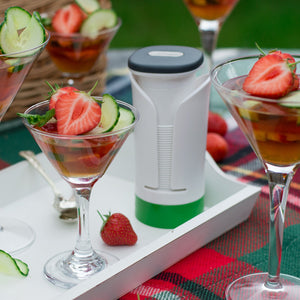 Four martini glasses with drinks garnished using spiral-cut strawberries, cucumber, and mint created with the Zyliss Spiralizer sit on a white tray atop a red plaid picnic blanket near a white insulated flask. 