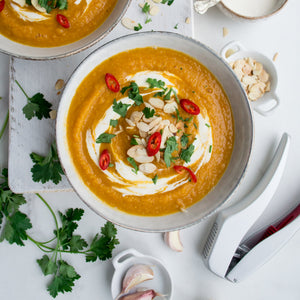 A bowl of orange soup with cream, chili, herbs, and almond flakes is surrounded by parsley, garlic, and the Zyliss Susi 3 Garlic Press featuring an ergonomic handle on a white surface. 