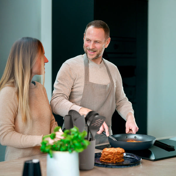 A man in an apron smiles while cooking pancakes at the stove with a heat-resistant Zyliss Spoon, chatting with a woman in their modern kitchen decorated with utensils and a potted plant on the counter. #Medium