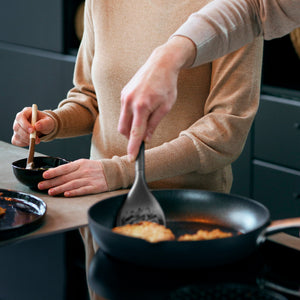Two people cook together; one stirs a small bowl, while the other uses the Zyliss Slotted Turner with an ergonomic handle to cook food in a frying pan on the stove. Their faces arent visible. 
